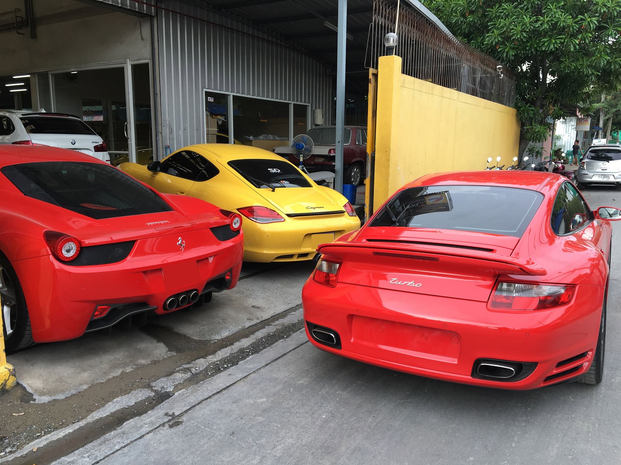 Ferrari 458 สีแดง Porsche Cayman สีเหลือง และ Porsche 911 Turbo สีแดงจอดหน้าศูนย์